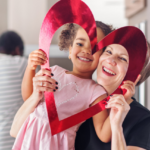 A joyful young girl and a middle-aged woman peek through a large, red heart-shaped cutout, both smiling, with another person visible in the background.