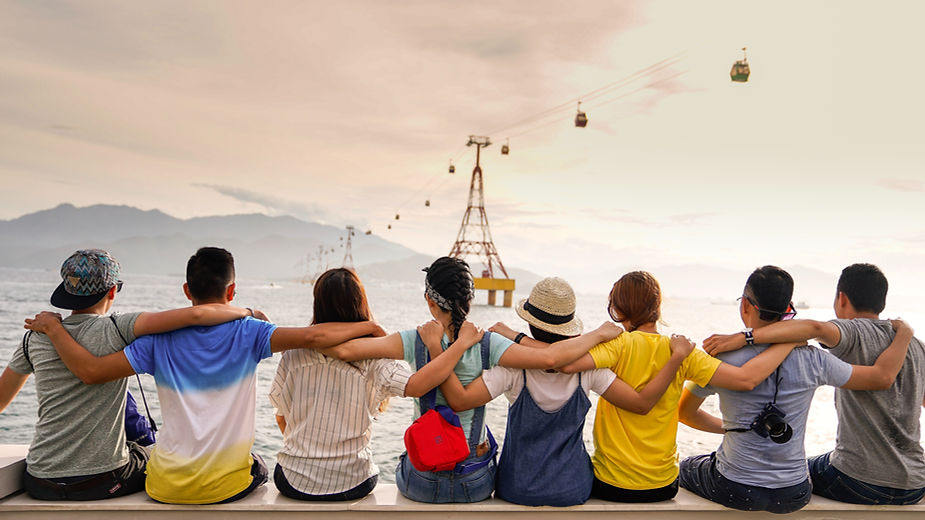 A group of seven friends sitting together by the waterfront, arms around each other's shoulders, watching cable cars move over the water at sunset.