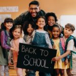 A joyful teacher surrounded by diverse group of young students holding a "back to school" chalkboard sign in a classroom setting.