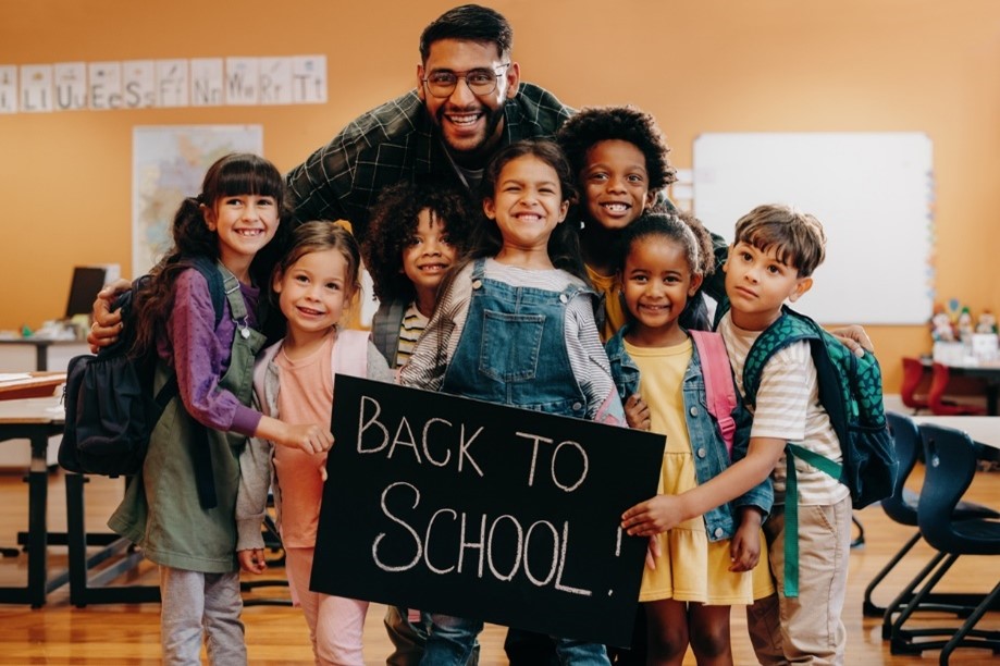 A joyful teacher surrounded by diverse group of young students holding a "back to school" chalkboard sign in a classroom setting.