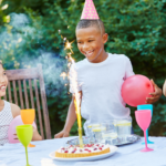 Three children wearing party hats having fun at a birthday party outdoors, with one boy lighting a sparkler on a cake as his friends look on with balloons and drinks.