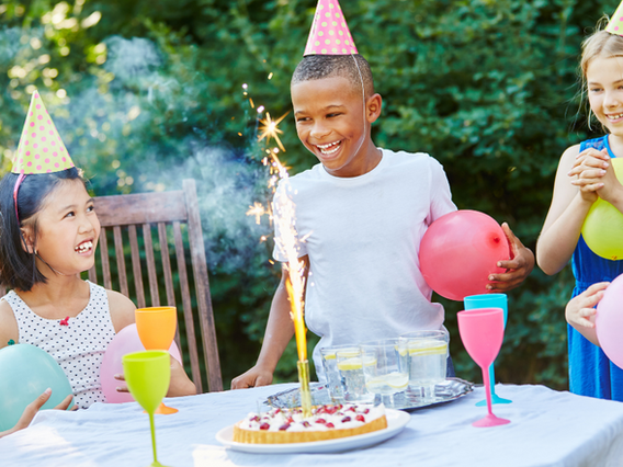 Three children wearing party hats having fun at a birthday party outdoors, with one boy lighting a sparkler on a cake as his friends look on with balloons and drinks.