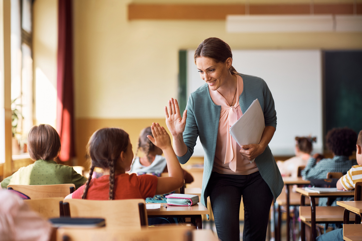 A female teacher is giving a high-five to a young girl in a classroom filled with students. the teacher is smiling and holding papers, while students sit at desks with books open.