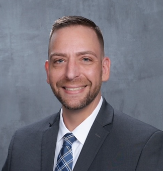 A professional headshot of a smiling man with short, styled hair and a beard, wearing a suit with a striped tie, against a gray background.