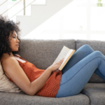 A woman with curly hair, wearing a red polka-dot top and jeans, is reading a book while reclining comfortably on a grey couch in a brightly lit room.