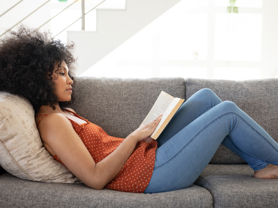 A woman with curly hair, wearing a red polka-dot top and jeans, is reading a book while reclining comfortably on a grey couch in a brightly lit room.