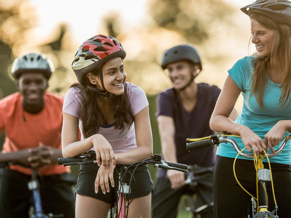 Four diverse friends enjoying a sunny day outdoors with their bicycles, smiling and conversing happily. one woman leans on her bike while chatting with another, both wearing helmets.
