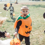 A joyful child in a pumpkin costume with a painted face celebrates halloween outdoors, holding a candy bucket, surrounded by other kids in costumes.