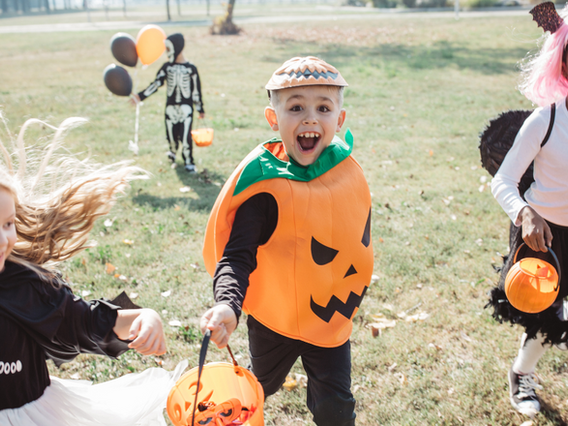 A joyful child in a pumpkin costume with a painted face celebrates halloween outdoors, holding a candy bucket, surrounded by other kids in costumes.