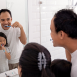A family engaging in their dental hygiene routine in the bathroom, with a father and his daughter brushing their teeth while smiling at each other in the mirror.