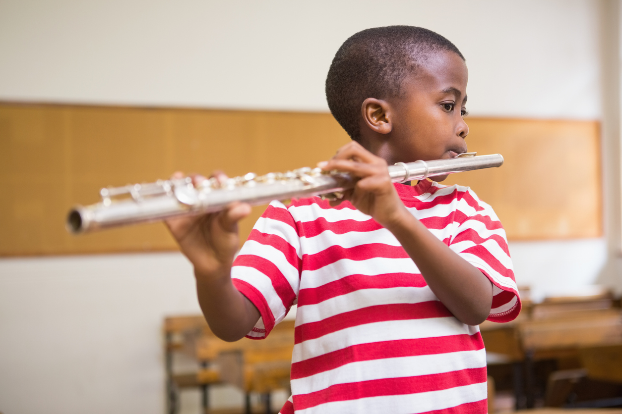 A young boy in a red and white striped shirt focused intently on playing a silver flute in a classroom setting.