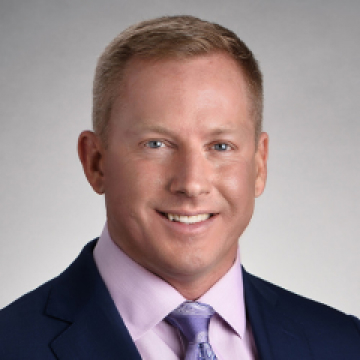 Professional headshot of a smiling man with short blonde hair, wearing a dark blue suit, light purple shirt, and purple tie against a neutral gray background.