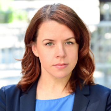 A professional headshot of a woman with shoulder-length auburn hair, wearing a blue blazer, and looking directly at the camera with a serious expression, against a blurred cityscape background.