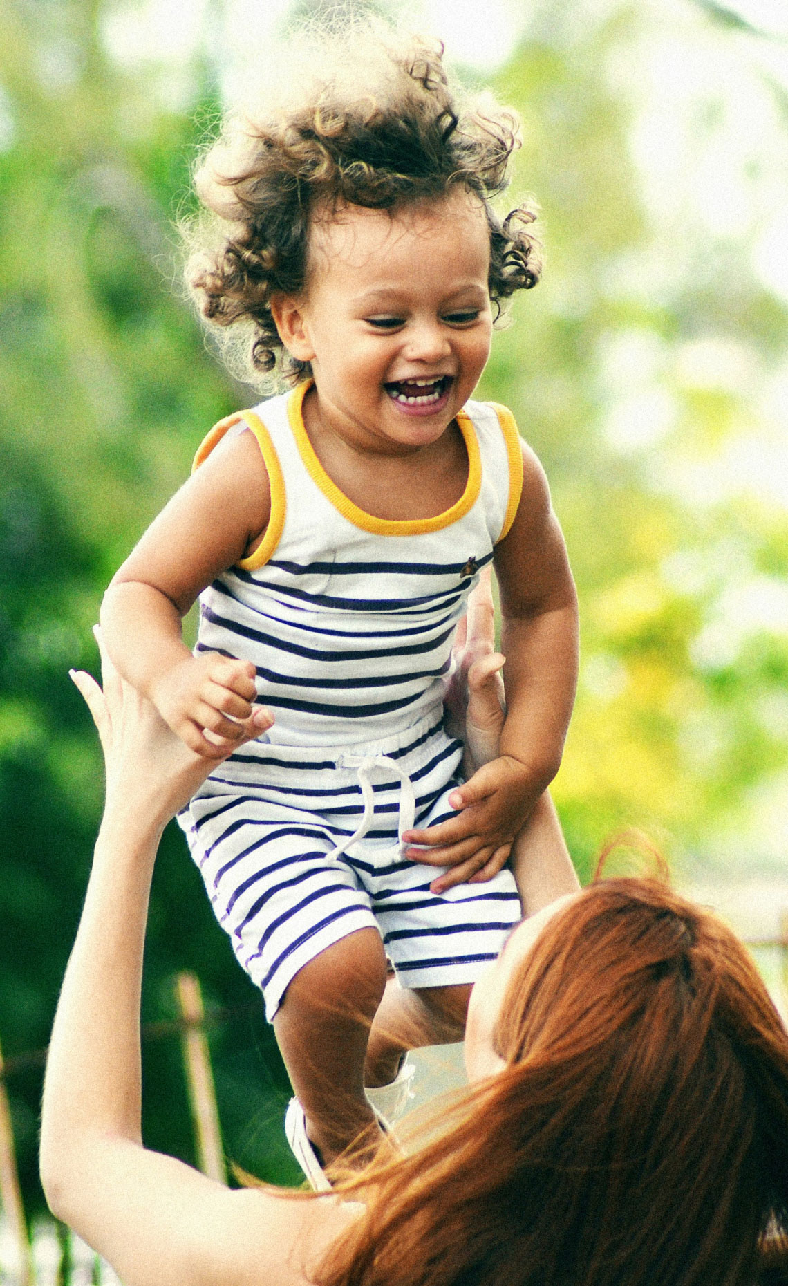 A joyful toddler with curly hair, dressed in a striped outfit, is being lifted high in the air by a woman with red hair, both outdoors surrounded by greenery.