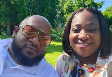 A smiling man and woman sitting outdoors on a sunny day, both wearing sunglasses, with trees and lush greenery in the background.