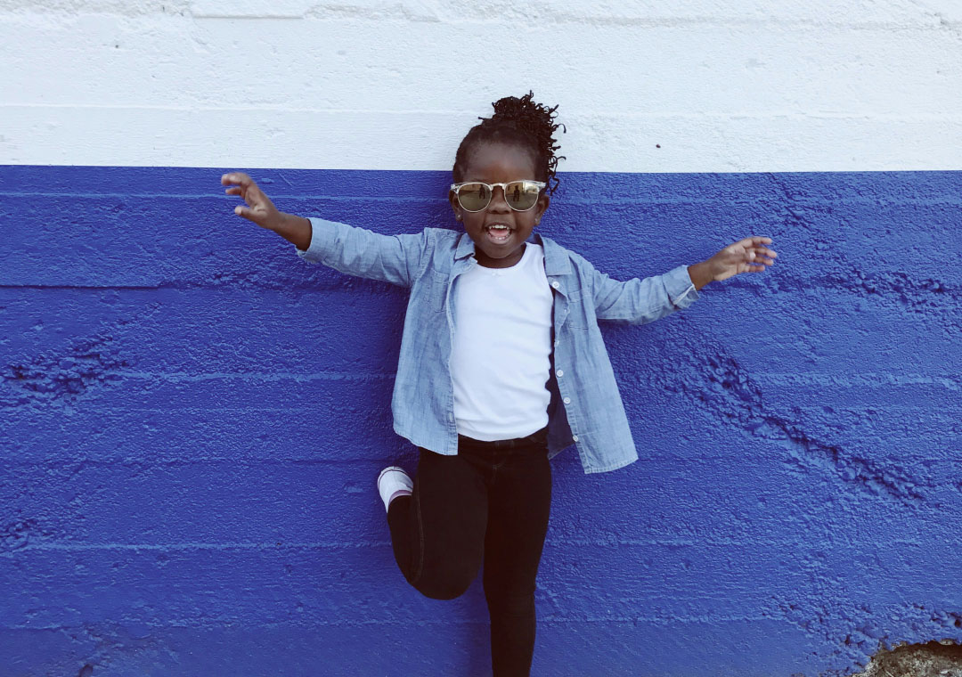 A joyful young girl with sunglasses and curly hair jumps in front of a blue wall, arms spread wide, wearing a white shirt, denim jacket, and black pants.