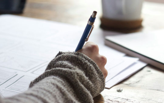 A person's hand wearing a knit sweater holds a pen over a paper on a wooden desk, with a coffee cup in the background.