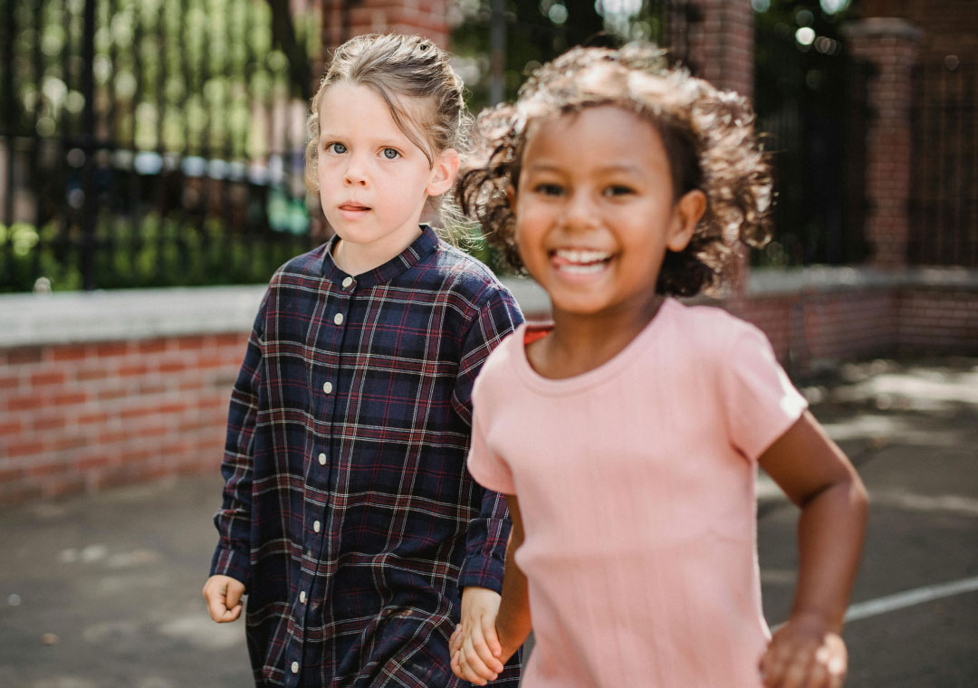 Two young children playing outside, one in a pink dress joyfully running towards the camera, and the other in a plaid shirt looking back with a focused expression.