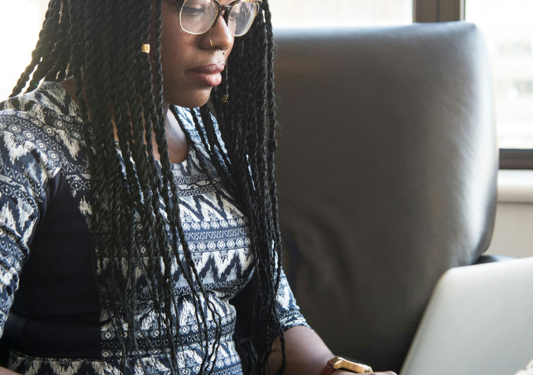 A woman with braided hair and glasses is focused on her work on a laptop in a bright office setting.