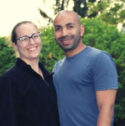 A smiling couple standing close together outdoors with trees and greenery in the background. the man is bald and wearing a blue t-shirt, and the woman is wearing glasses and a black jacket.