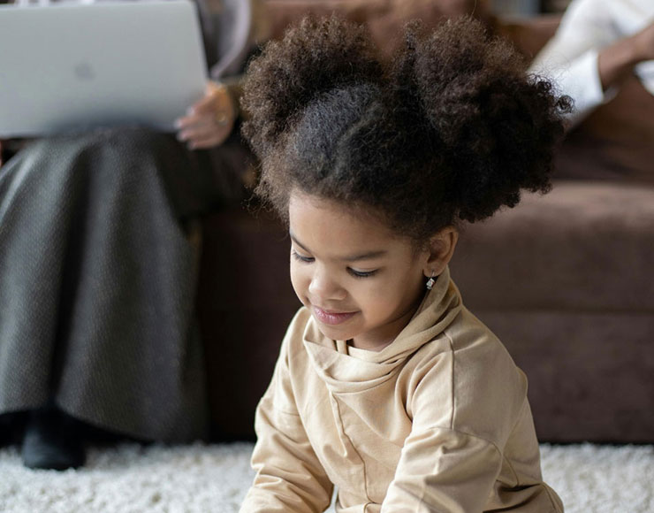 A young girl with two puffy ponytails is focused on a digital tablet, sitting on the floor. in the background, an adult's legs are visible beside a laptop on a couch.