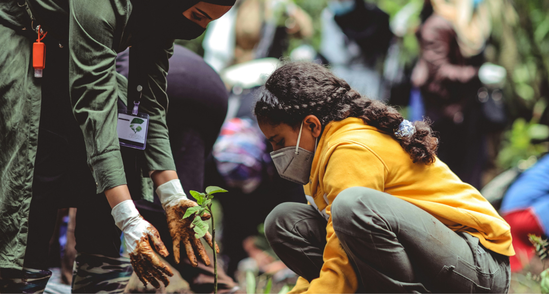 A group of people participate in a tree planting event, focusing on a young woman in a yellow hoodie and mask planting a sapling.