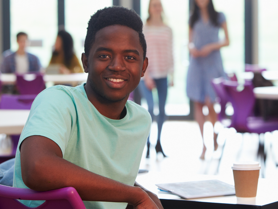 A young man smiling at the camera while sitting in a busy college cafeteria, with other students in the background. he is wearing a light teal t-shirt and seated at a purple desk.