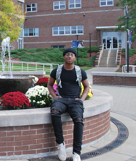 A young man in a black outfit and cap sits relaxed on a brick wall, with a backpack, in front of a building and a fountain surrounded by colorful flowers.