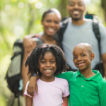 A happy family with two children, smiling in a sunny forest setting, enjoying a day of hiking. the parents are in the background, and the children are in front.