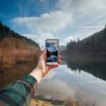 A person's hand holding a smartphone capturing a photo of a serene lake surrounded by forested hills under a partly cloudy sky.