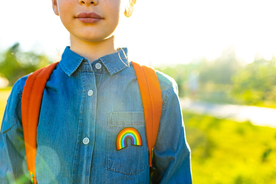 A child in a denim shirt with a rainbow patch and an orange backpack stands outdoors in bright sunlight, with a blurred green background.