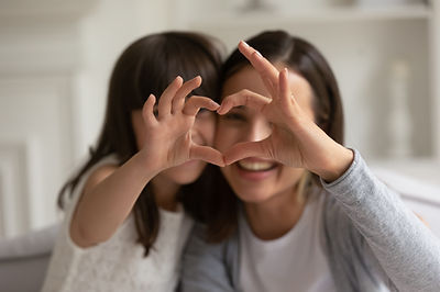A joyful woman and a young girl sitting closely, smiling as the woman creates a heart shape with her fingers in front of her eye. they are in a bright indoor setting.