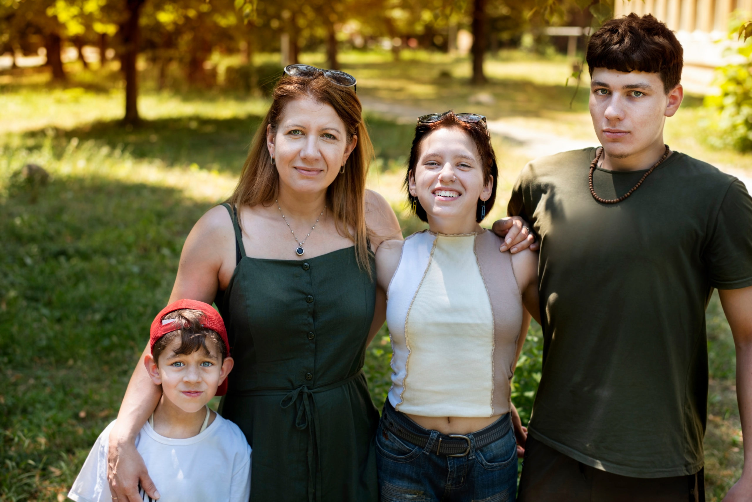 A family of four, including a mother and three children of varying ages, stands closely together in a sunny park, smiling at the camera. the mother and her two teenagers appear, while the youngest child wears a red cap.