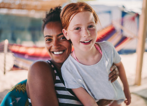 A smiling woman with dark skin embracing a joyful young girl with red hair, both looking at the camera, with a beach and hammocks blurred in the background.