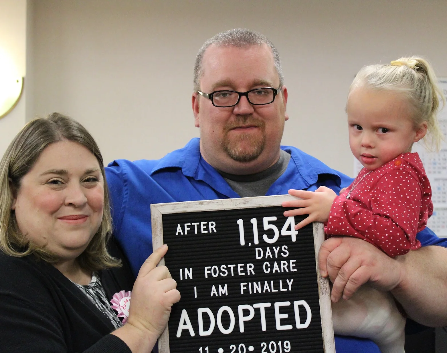 Two foster parents with adopted foster child with a sign that reads "after 1,154 days in foster care, I am finally adopted."