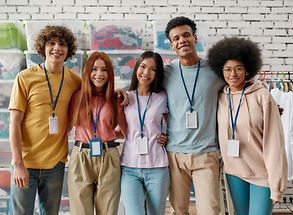 Five young adults with diverse backgrounds, wearing casual clothing and lanyards, smiling and standing together in a brightly-lit room with a brick wall background.