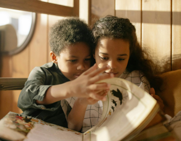Two children, a boy and a girl, are engrossed in a storybook. the boy, pointing at the page, shares something exciting with the girl as they read together in a warmly lit room.