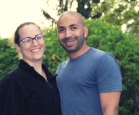 A smiling couple standing together outdoors, with a background of blurred green foliage. the woman wears glasses and a black top, and the man is in a blue shirt, bald.