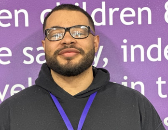 A man with glasses and a beard smiles gently, standing in front of a purple backdrop with text, wearing a black hoodie and a blue lanyard.