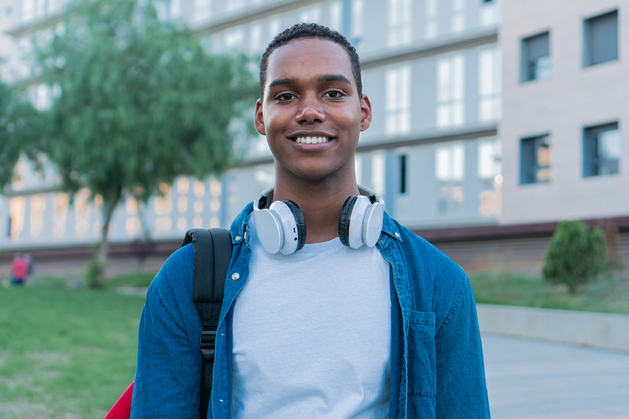 A young man smiles at the camera while wearing headphones around his neck and carrying a backpack. He stands outdoors in front of a modern building with a green space and a tree to his left. He is dressed in a blue shirt and a white t-shirt.