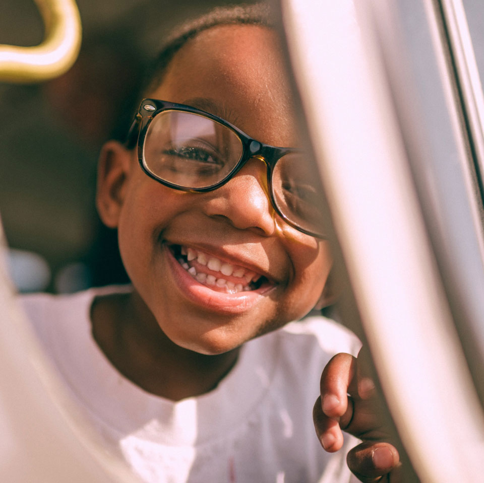 A joyful young boy wearing glasses smiles broadly, peeking through a gap with sunlight illuminating his face.
