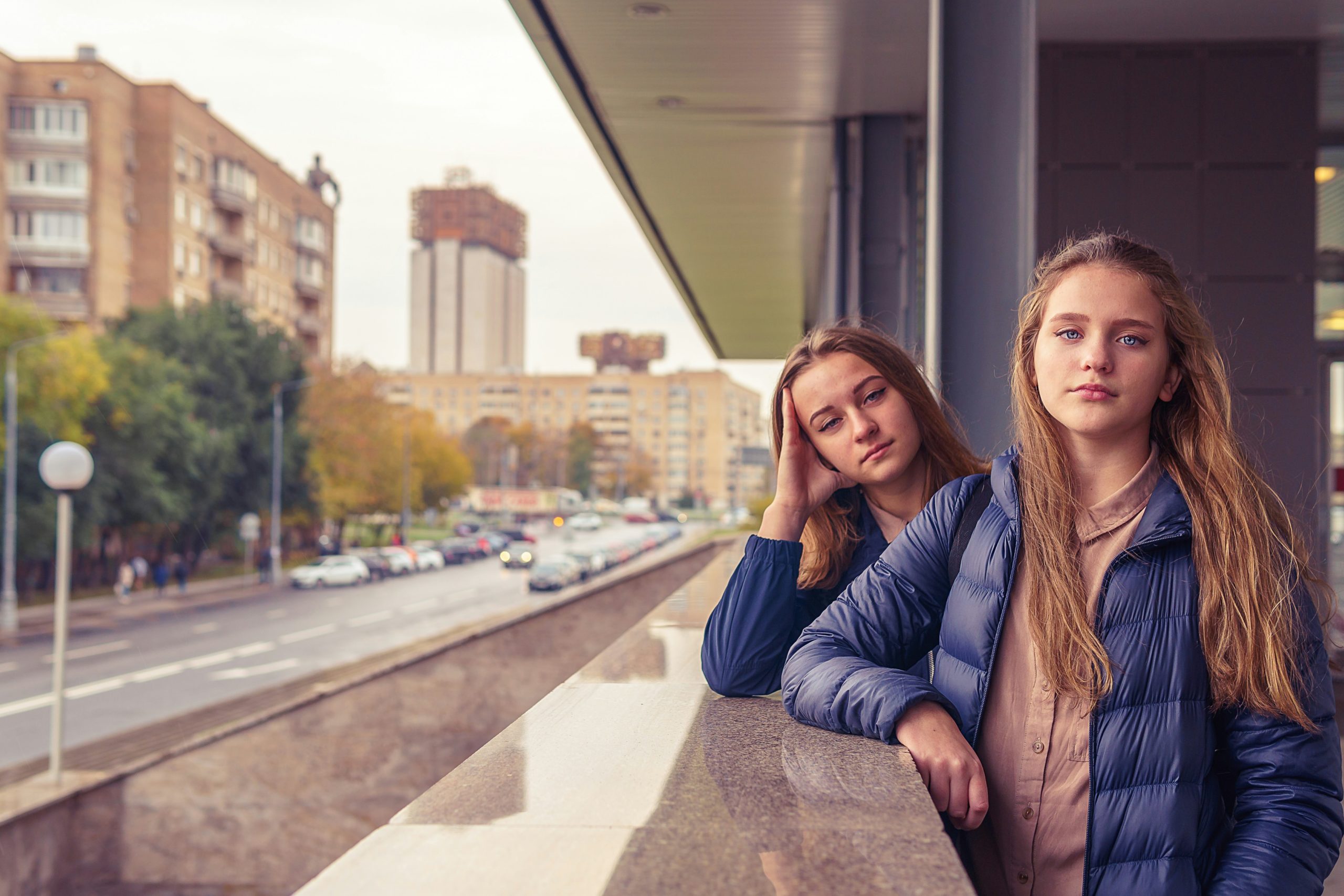Two young women in blue jackets leaning on a railing, looking thoughtful, with a city street and buildings in the background during the evening.