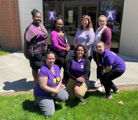 A group of seven women posing outdoors in front of a building with purple ribbons and a logo on the door. they wear various shades of purple clothing, smiling at the camera.