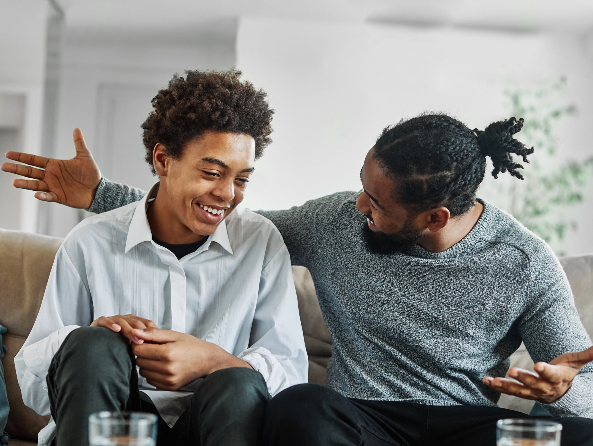 Two black males, one youth and one adult, sitting closely on a couch, engaged in a joyful conversation.