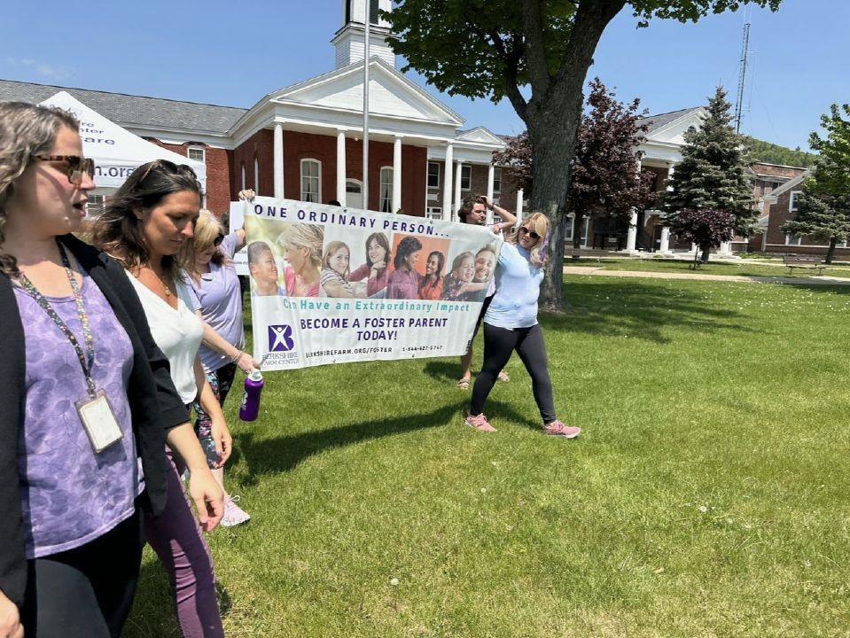 A group of people walking on grass in front of a building. They are holding a banner that reads, "ONE ORDINARY PERSON... CAN HAVE AN EXTRAORDINARY IMPACT. BECOME A FOSTER PARENT TODAY!" with images of smiling individuals and a logo for a Berkshire Farm.