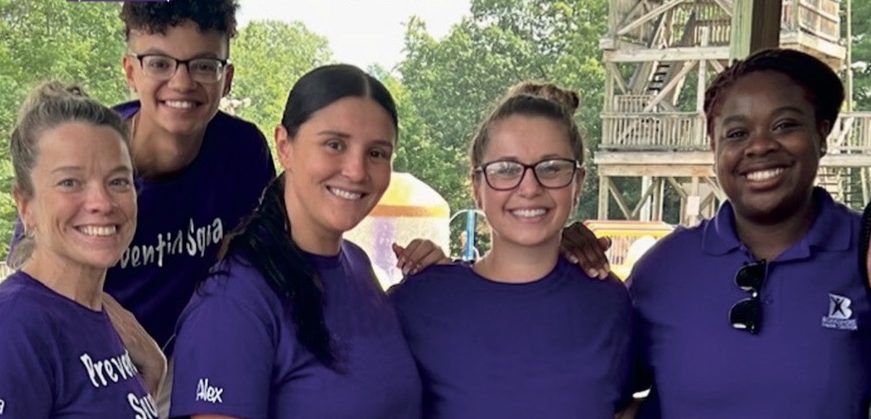 A diverse group of five people wearing purple shirts and smiling, standing under a wooden structure. The text 