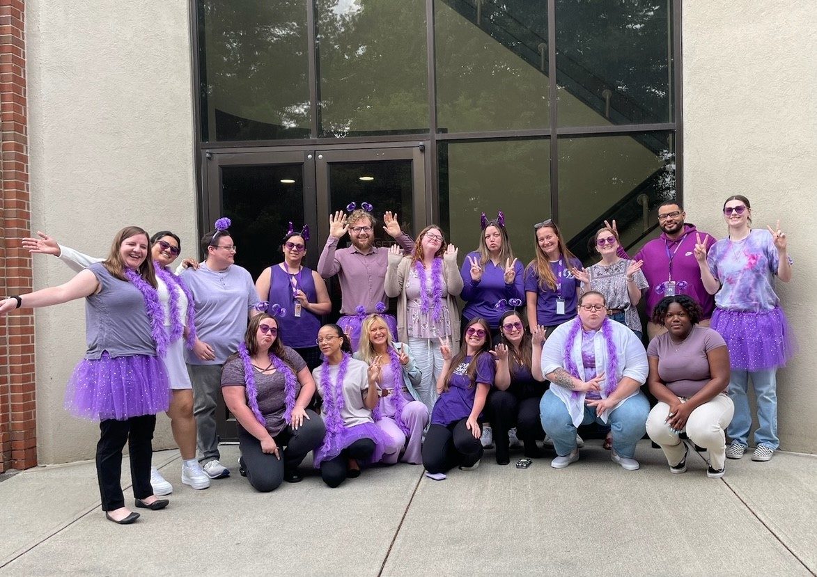 A group of people stand and crouch in front of a building. They are dressed in various shades of purple, some wearing accessories like tutus, headbands, and scarves. Everyone is smiling, waving, or posing cheerfully. The setting appears to be a casual event.