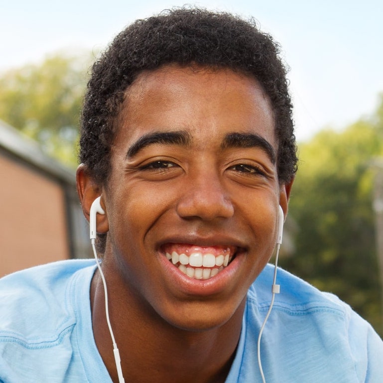 A teen boy with short curly hair and wearing earphones smiles brightly at the camera. He is dressed in a light blue shirt and is outdoors, with a blurred background of trees and buildings.