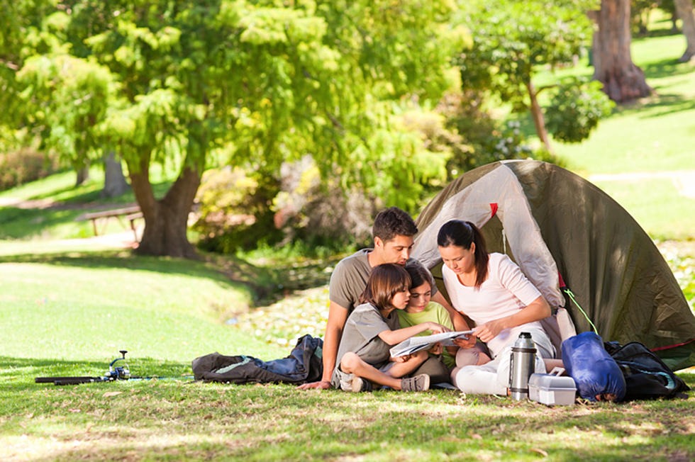 A family of four, including two children and their parents, sits outside a pitched tent in a park. They are gathered closely, looking at a map together. The park is vibrant with greenery and sunlight, creating a serene atmosphere. Camping gear is scattered around.