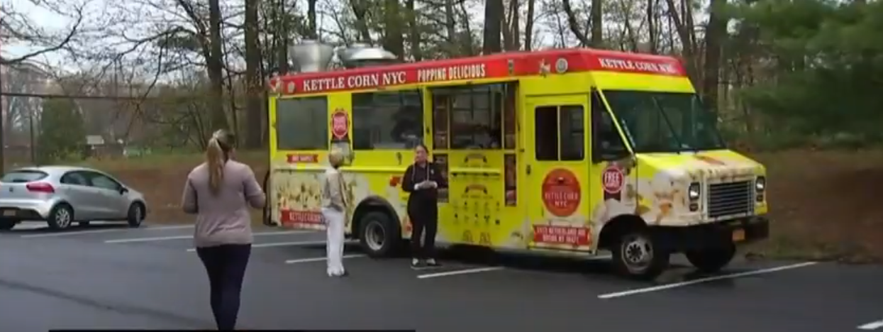 A yellow food truck labeled "Kettle Corn NYC" is parked in a lot with trees in the background. Two people are standing near the truck, and one person is walking away. Another car is parked nearby. The scene appears to be in a casual, outdoor setting.